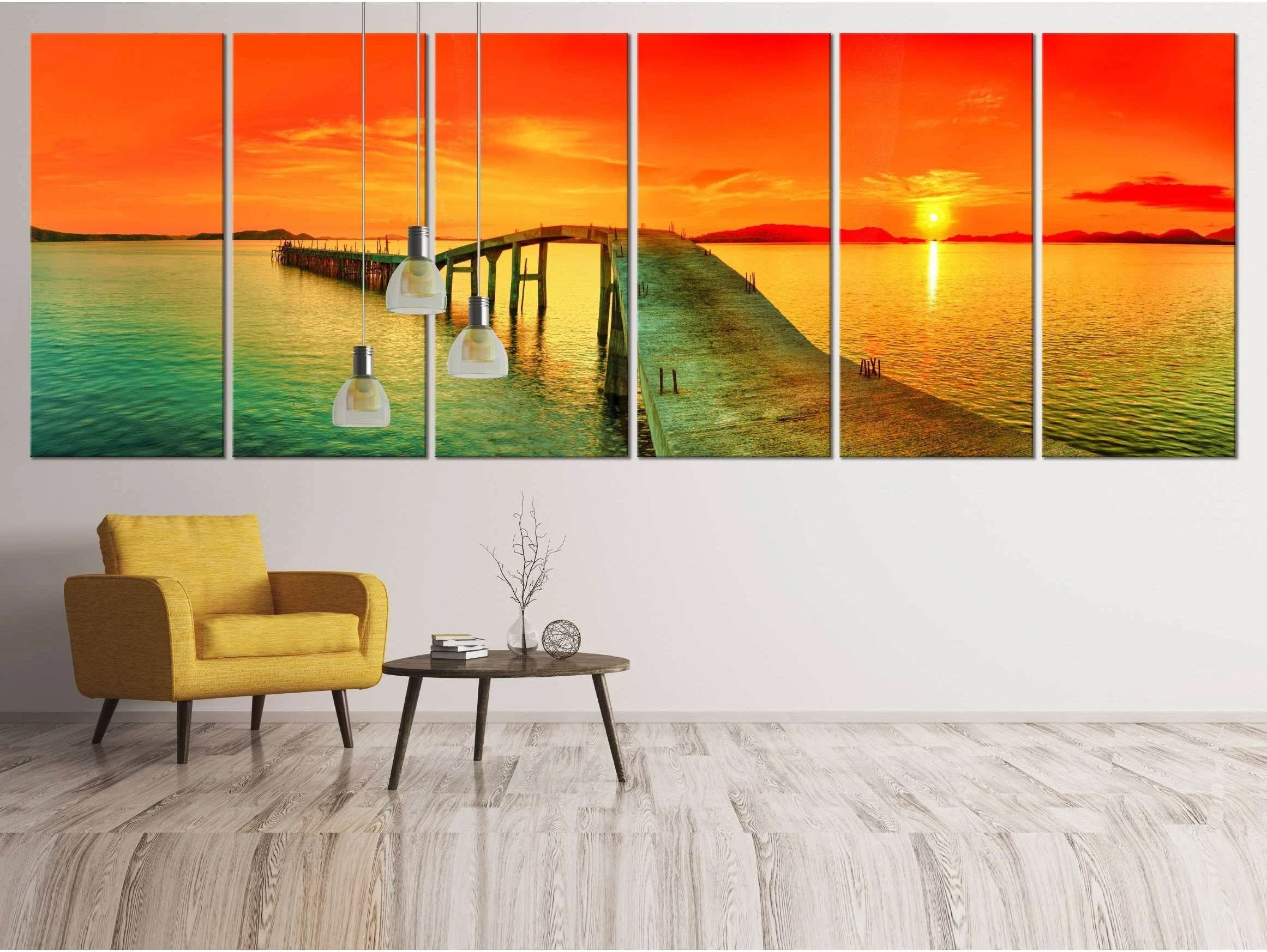 Multi-panel sunset pier canvas print above a chair in a modern living room with orange sky and teal sea